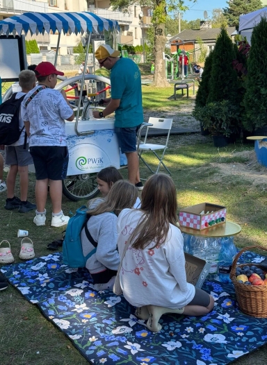 Grupa dzieci na pikniku w parku, trzy siedzą na kocu obok kosza z owocami, a dwie stoją przy stoisku z napojami obsługiwanym przez mężczyznę.