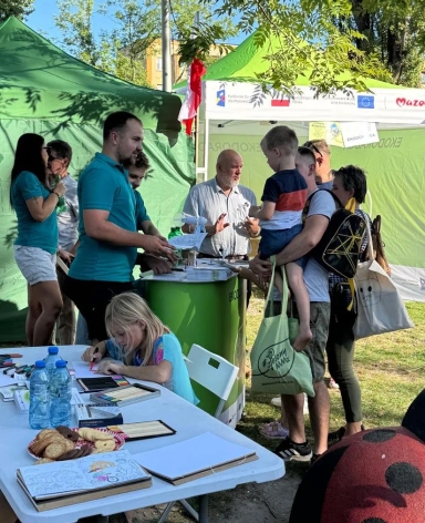 Grupa ludzi w plenerze przy stoiskach z zielonymi namiotami, dziecko rysujące przy białym stole z kredkami i wodą, obok mężczyzna rozmawiający z rodziną, rodzice trzymający dziecko na rękach.