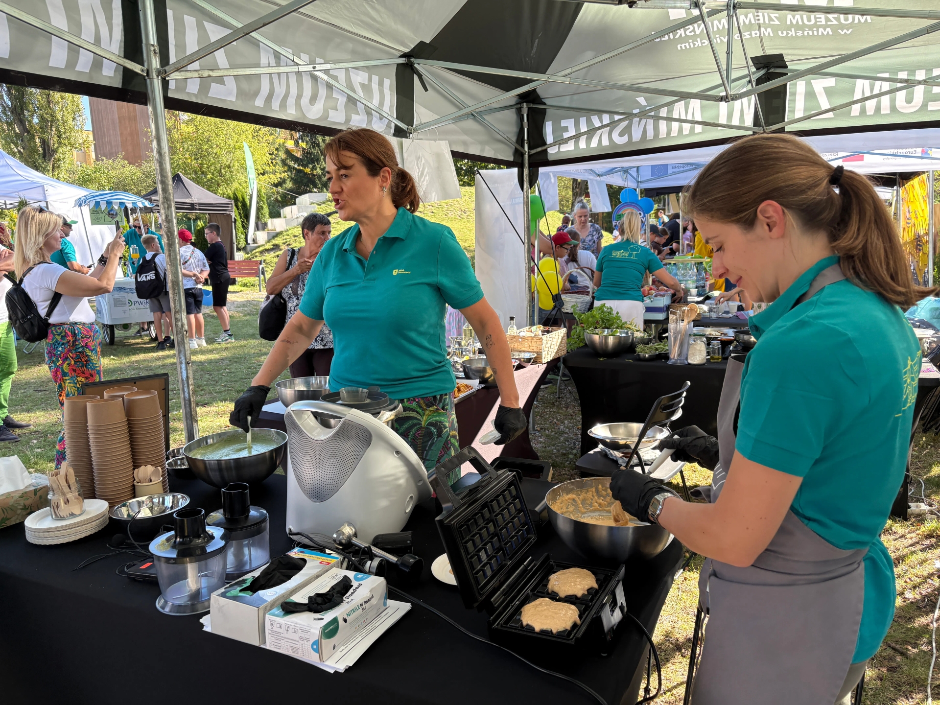 Pod namiotem kuchennym dwie kobiety w turkusowych koszulkach przygotowują potrawy na zewnątrz w ciągu dnia, w tle ludzie i inne namioty.