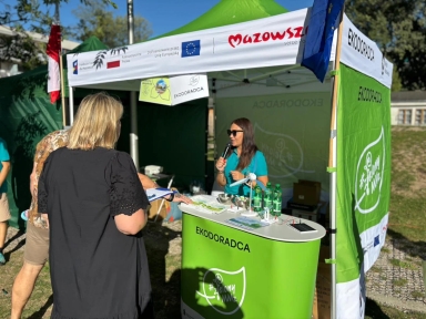 Kobieta w turkusowej koszulce i okularach przeciwsłonecznych stoi za zielonym stoiskem z napisem EKODORADCA i rozmawia z dwiema osobami. Stoisko znajduje się pod namiotem z banerem Mazowsze i flagą Unii Europejskiej. Na stole stoją butelki z wodą i model turbiny wiatrowej. W tle zieleń i niebieskie niebo.