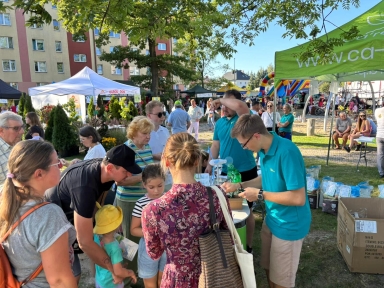 Grupa ludzi na zewnątrz przy stoisku z zielonym namiotem i plakatami, w słoneczny dzień na festynie, pośród innych namiotów i budynków mieszkalnych w tle.