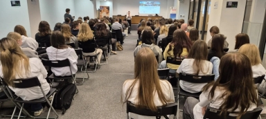 Grupa młodych osób siedzi na krzesłach w sali konferencyjnej, skierowanych w stronę kobiety przemawiającej na podwyższeniu z rzutnikiem na ścianie. Sala ma jasne ściany i szarą wykładzinę.