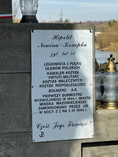 Biała metalowa tablica pamiątkowa zamocowana na szarej kamiennej ścianie. Tablica ma czarny nadruk i lekkie pęknięcia powierzchni. Po prawej stronie stoją kilka przezroczystych szklanych zniczy ze złotymi przykrywkami. Na górze po lewej widać wysoki szklany lampion. W tle widoczne jezioro, drzewa i lekko zachmurzone niebo w świetle dziennym.