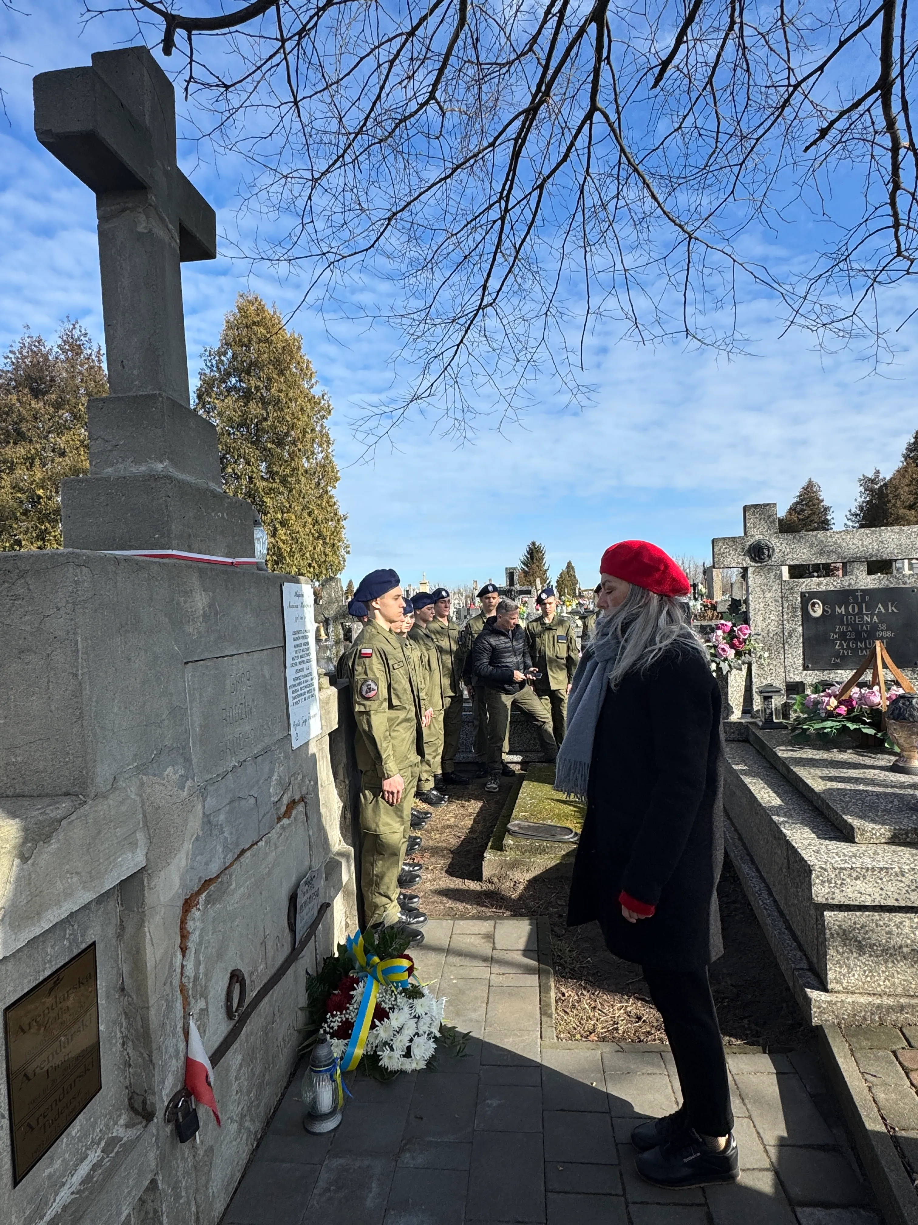 Duży kamienny krzyż stoi po lewej stronie na cmentarzu. Rząd kilkunastu żołnierzy w mundurach stoi naprzeciw pomnika. Po prawej stoi kobieta w czerwonym berecie i szarym szalu. U podstawy pomnika leży wieniec z wstążką w kolorach niebieskim i żółtym oraz znicz. Na niebie widoczne są gołe gałęzie drzew i błękitne chmury.