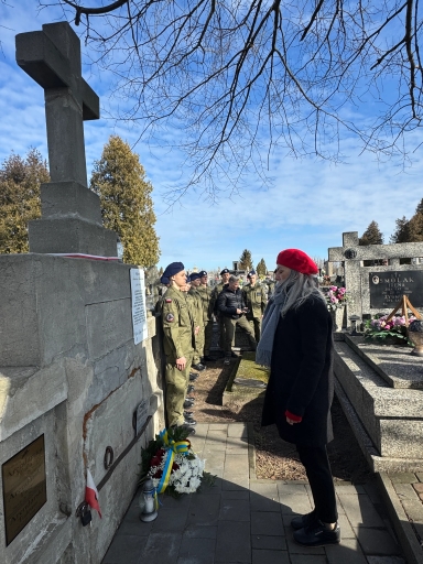 Duży kamienny krzyż stoi po lewej stronie na cmentarzu. Rząd kilkunastu żołnierzy w mundurach stoi naprzeciw pomnika. Po prawej stoi kobieta w czerwonym berecie i szarym szalu. U podstawy pomnika leży wieniec z wstążką w kolorach niebieskim i żółtym oraz znicz. Na niebie widoczne są gołe gałęzie drzew i błękitne chmury.