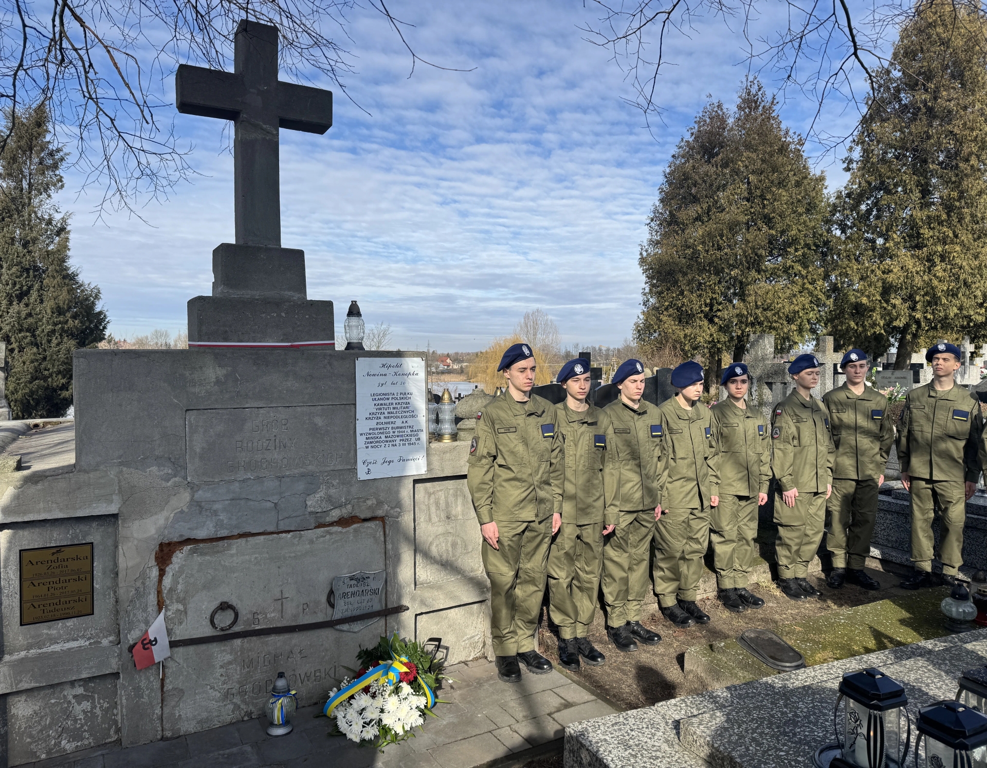 Duży kamienny krzyż stoi po lewej nad nagrobkiem. Dziesięć umundurowanych osób w beretach stoi w szeregu po prawej. Na nagrobku leży biały wieniec z kolorową wstążką. Otoczenie cmentarza z drzewami i niebem w tle w dzień.