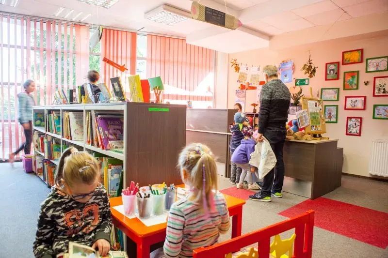 Dzieci siedzące przy małych stolikach z przyborami do rysowania oraz półki z książkami i dorosły stojący przy ladzie w jasnym pokoju z wiszącymi rysunkami na ścianie.