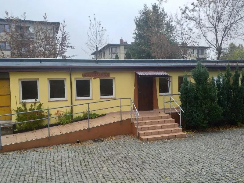 Żółty budynek z czterema oknami, wejściem z drewnianymi drzwiami, podjazdem z poręczą i schodami, otoczony krzewami.