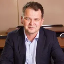 Mężczyzna w marynarce i jasnej koszuli siedzi przed biurkiem.