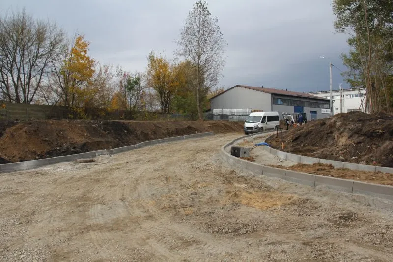 Budowa drogi z widocznym korytem i krawężnikami, biała furgonetka oraz budynki w tle.