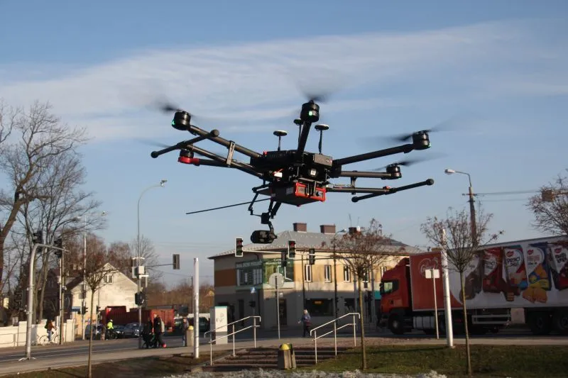 Czarny wielowirnikowy dron lata nad ulicą w miejskim otoczeniu z budynkami i pojazdami.