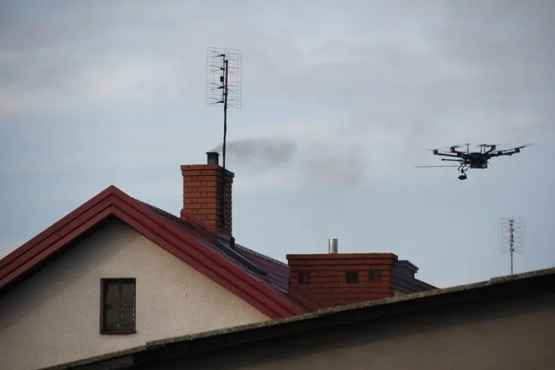 Dach budynku z czerwoną dachówką, komin z dymem i latający dron w powietrzu.