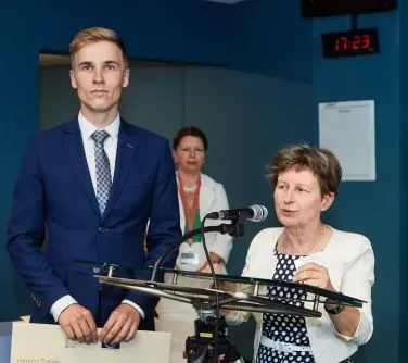 Mężczyzna w garniturie stoi obok kobiety w białej marynarce mówiącej do mikrofonu przy pulpicie, a w tle znajduje się kolejna kobieta.