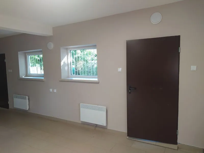 Ściana z dwoma oknami z kratami, dwoma grzejnikami oraz ciemnymi drzwiami z klamką.