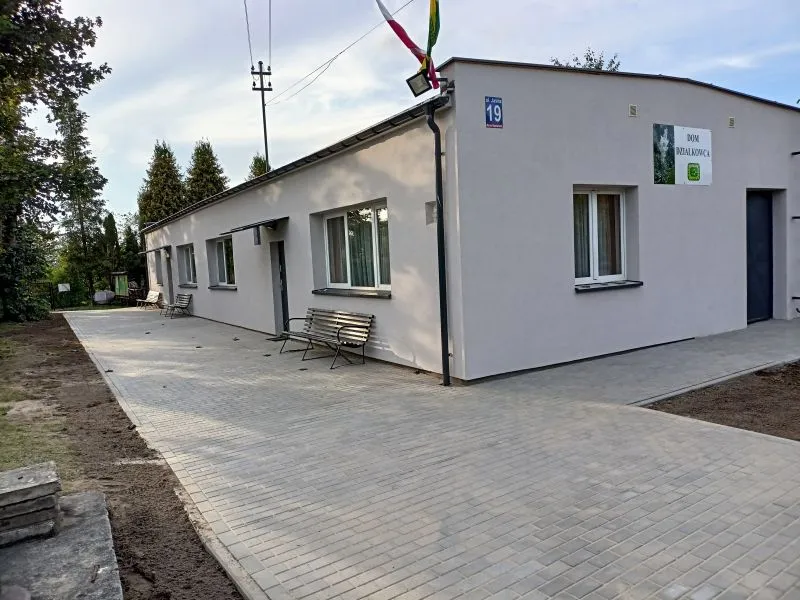 Jeden parterowy budynek z białą elewacją, kilkoma oknami, ławkami przed wejściem oraz tablicą z napisem „Dom Działkowca”.