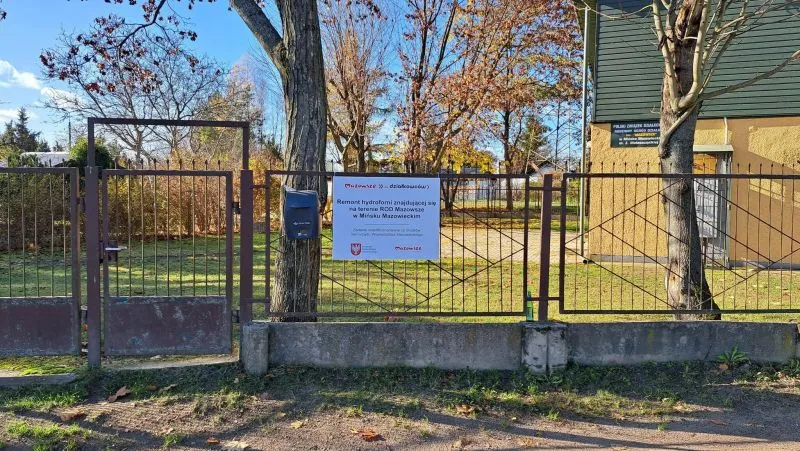 Metalowy płot z bramą i tablicą informacyjną o remoncie hydroforni na tle trawnika i drzew.