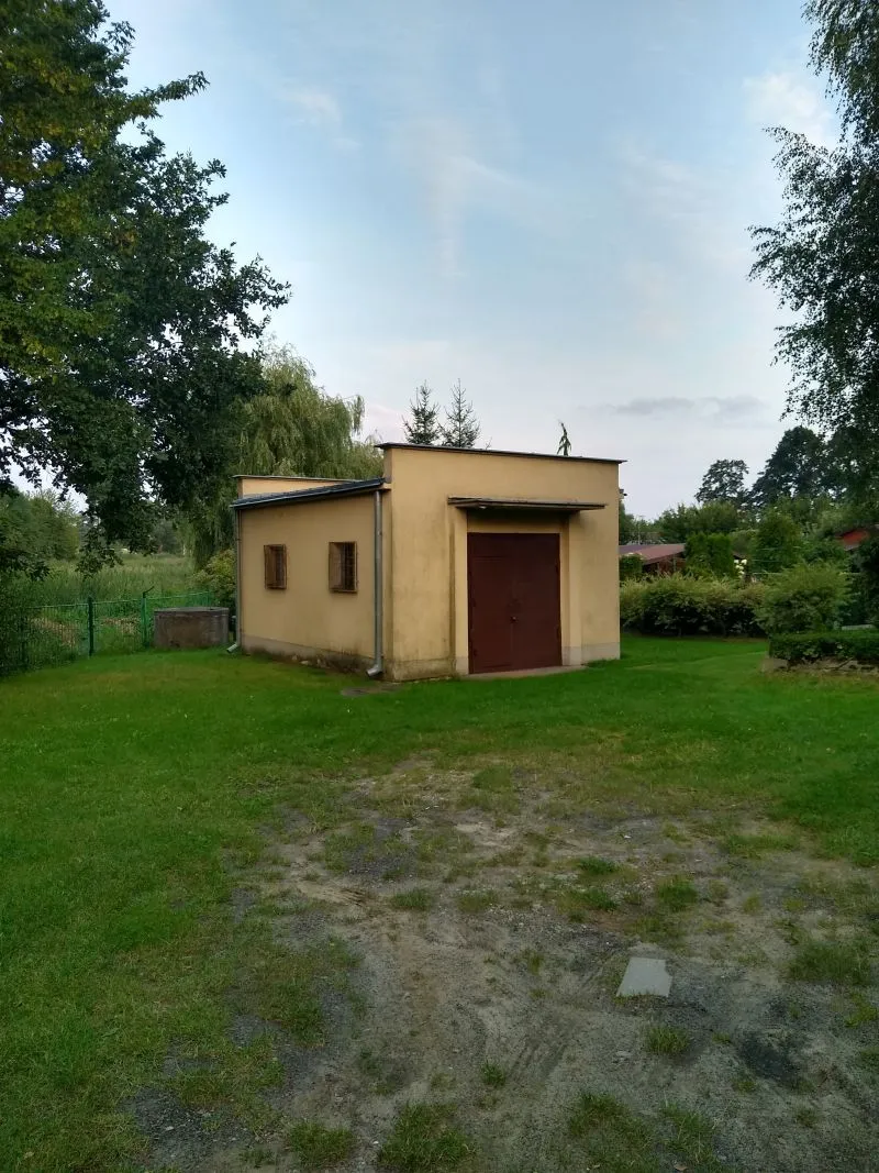 Prostokątna, żółta budka z brązowymi drzwiami i dwoma małymi oknami na zielonym trawniku otoczona drzewami.