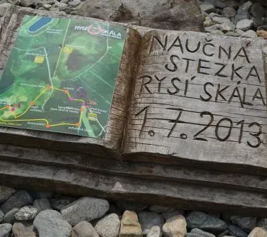 Drewniana tablica z mapą ścieżki edukacyjnej i napisem „Naučná stezka Rysí skála 1.7.2013” leżąca na kamieniach.