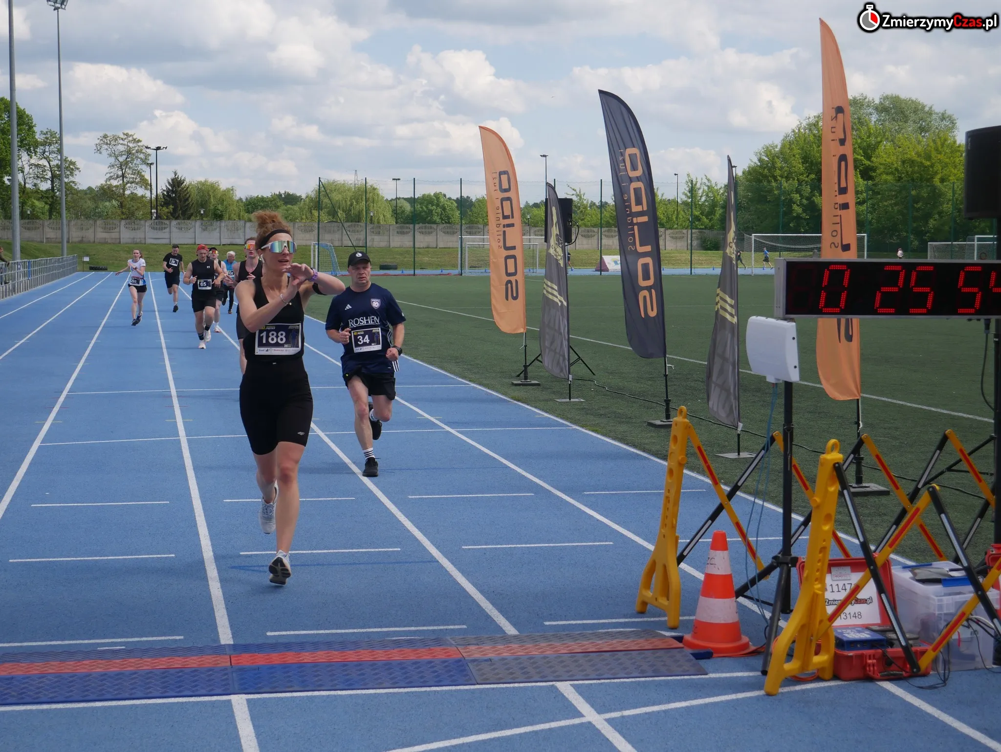Biegacze przekraczający metę na niebieskiej bieżni z widocznym czasem 25:54 na elektronicznym liczniku.
