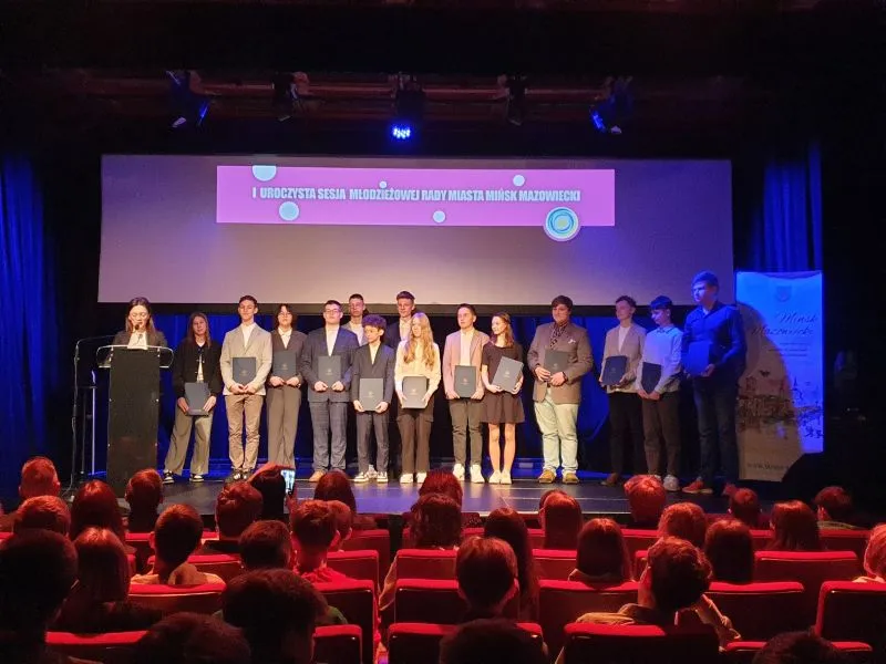 Grupa młodych osób stoi na scenie w rzędzie, trzymając teczki, obok stoi osoba przy mównicy, a przed nimi siedzi publiczność.