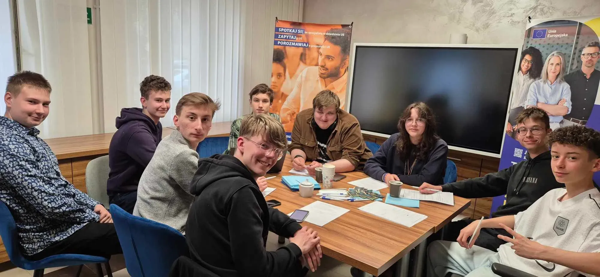 Dziewięć młodych osób siedzi przy stole z dokumentami, długopisami i kubkami, na tle dużego ekranu oraz dwóch stojących plakatów.