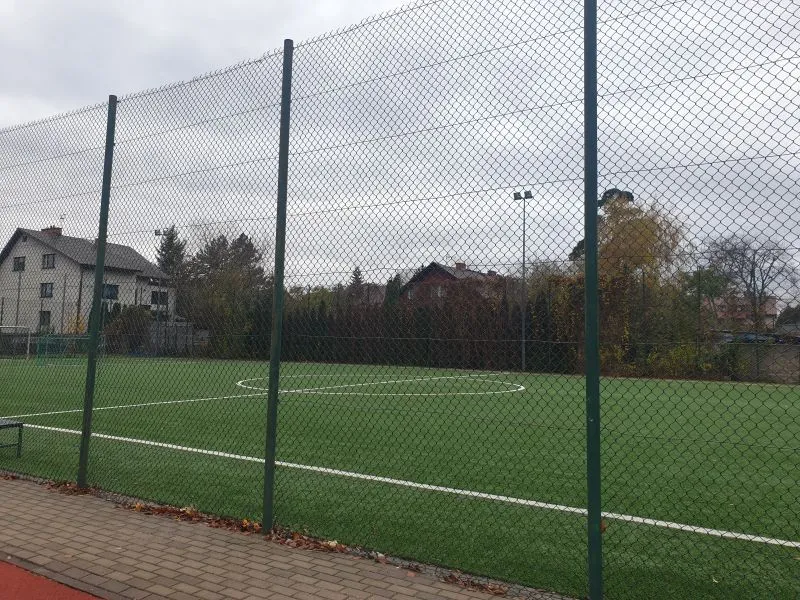 Orlik z zieloną murawą otoczony siatką i słupami, obok alejka z kostki brukowej.