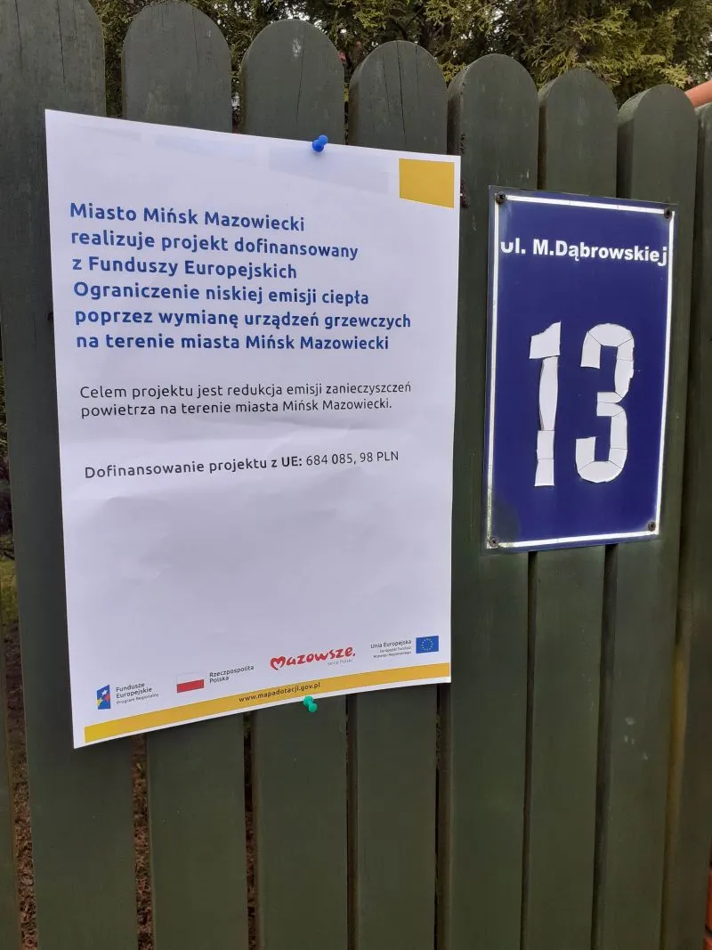 Tabliczka z numerem 13 i plakat informujący o projekcie dofinansowanym z funduszy europejskich na ograniczenie niskiej emisji ciepła w Mińsku Mazowieckim, umieszczone na zielonym drewnianym płocie.