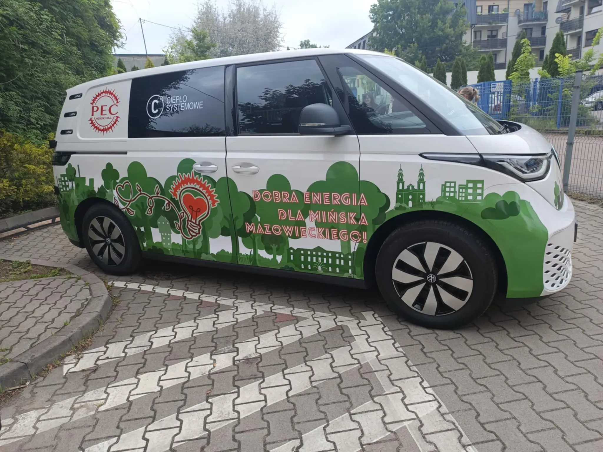 Biały van z zielonym motywem drzew, budynków oraz napisami „PEC Mińsk Maz.”, „Ciepło systemowe” i „Dobra energia dla Mińska Mazowieckiego!” zaparkowany na chodniku.