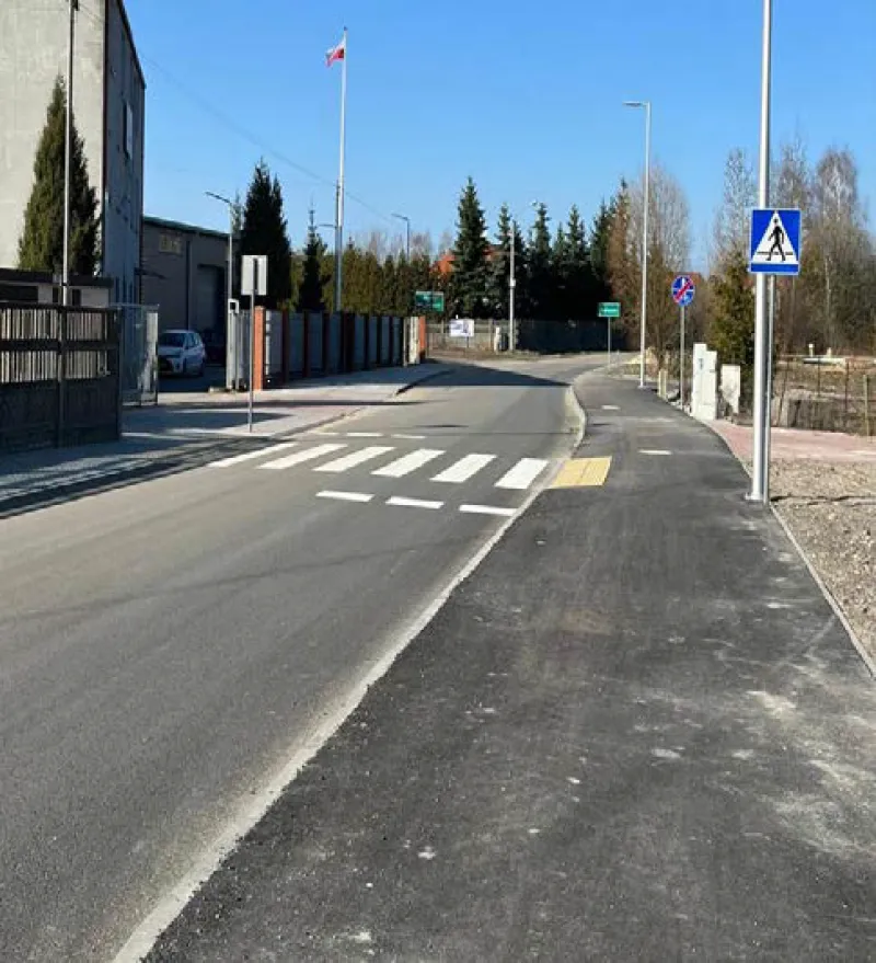 Ulica z przejściem dla pieszych, chodnikiem, znakami drogowymi i zabudowaniami po lewej stronie.