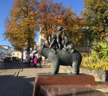 Rzeźba przedstawiająca niedźwiedzia z trzema dziećmi siedzącymi na jego grzbiecie, ustawiona na cokole na chodniku.