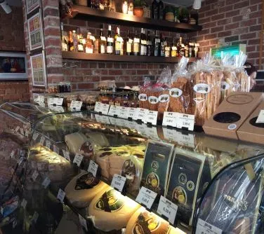 Lada sklepowa ze szklanym witryną zawierająca sery, przekąski i słoiki oraz półki z butelkami alkoholu i opakowaniami przekąsek.